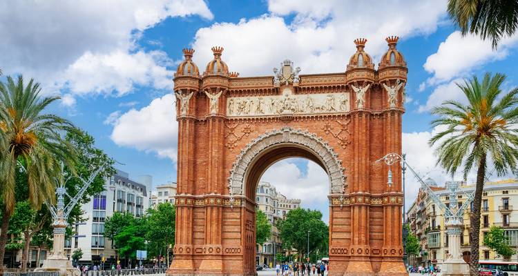 Rode bakstenen Arc de Triomf in Barcelona omlijst door palmbomen en enkele voetgangers onder een heldere hemel.
