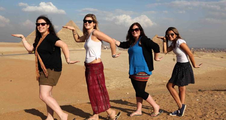 Four young women pose playfully in front of a distant pyramid on a sandy plateau.