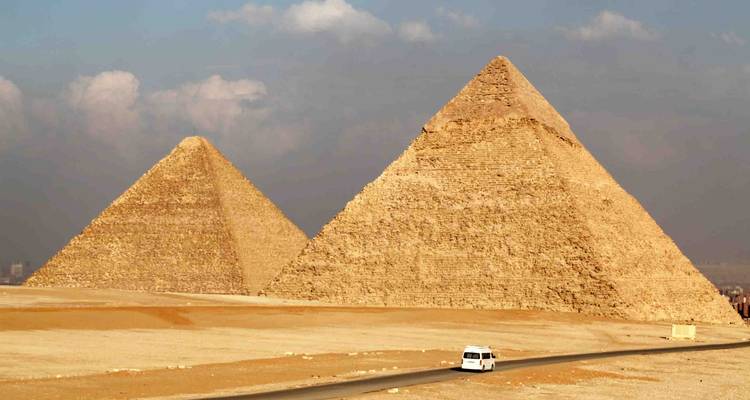 A clear view of two large pyramids rising from a flat sandy expanse with a lone van on a road.
