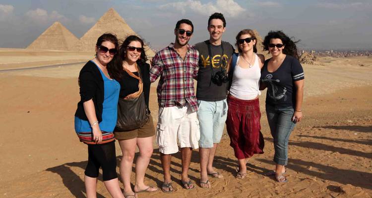 Six smiling travellers stand arm-in-arm on desert sand with pyramids in the background.