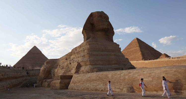 The Great Sphinx with two pyramids under a blue sky while several visitors walk nearby.
