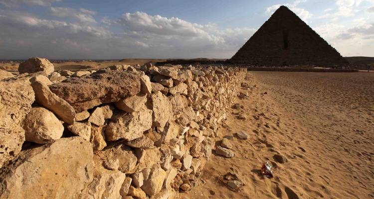 A stone wall leads the eye toward a shadowed pyramid across a sandy, litter-specked terrain.