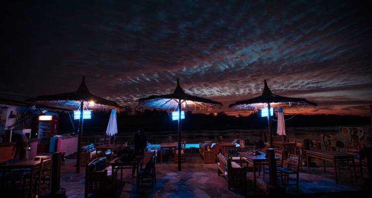 Open-air beach café at dusk with straw parasols silhouetted against a dramatic twilight sky.