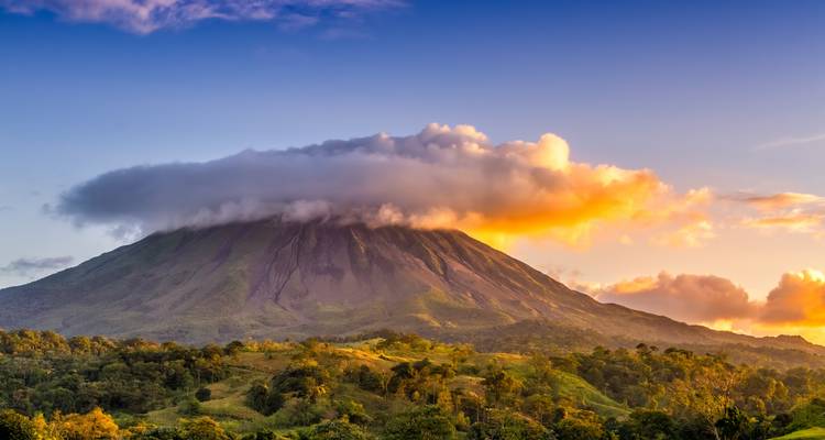 Wolkengekrönter kegelförmiger Vulkan, der sich über üppigen grünen Wald in warmem goldenem Licht erhebt.