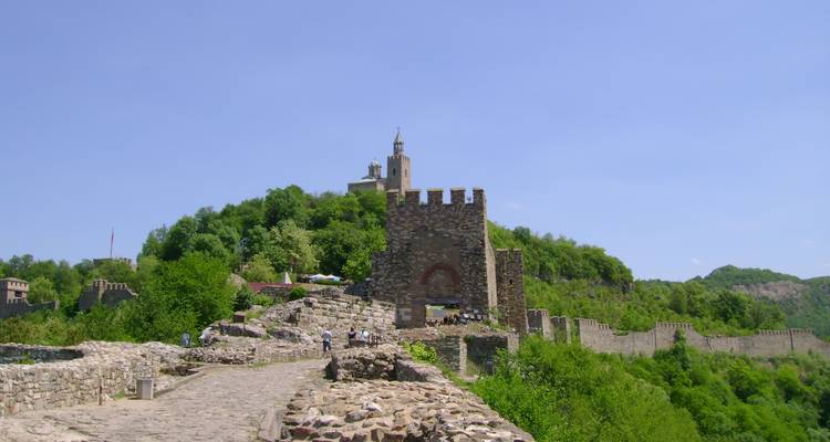 Des remparts de pierre et la porte de la forteresse de Tsarevets mènent à une église au sommet d'une colline sous un ciel bleu clair.