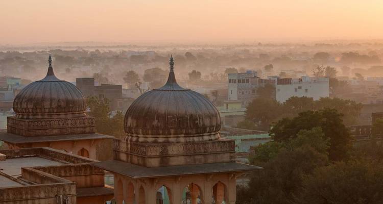 Sanftes Sonnenlicht der Morgendämmerung umhüllt zwei verzierte Kuppelpavillons mit Blick auf eine diesige Stadtlandschaft Rajasthans.