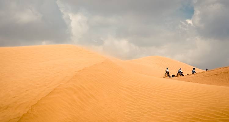 Petite caravane avec des cavaliers traversant des dunes ondulantes sous un ciel partiellement nuageux.