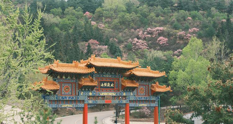 Ein farbenfrohes, mehrstöckiges chinesisches Tor überspannt eine gewundene Bergstraße inmitten grüner Kiefern und rosa blühender Bäume.