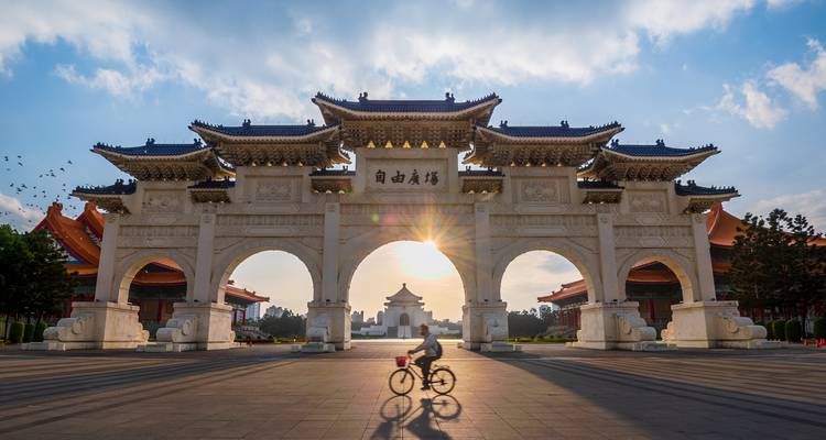 Gebogen Liberty Square Poort omlijst een gloeiende zonsopgang met een fietser als silhouet voor Taipei's Chiang Kai-shek Memorial Hall.