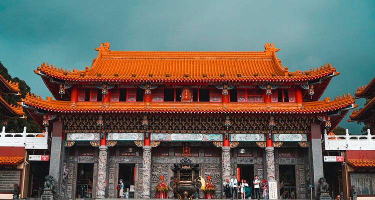 Templo de techo rojo brillante con fachada ricamente decorada se alza contra un cielo tormentoso color verde azulado mientras los visitantes se reúnen abajo.