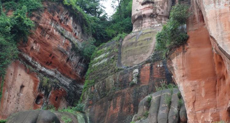 Gros plan sur le torse massif sculpté, la main et le visage à flanc de falaise du Bouddha géant de Leshan entourés de roche rouge.