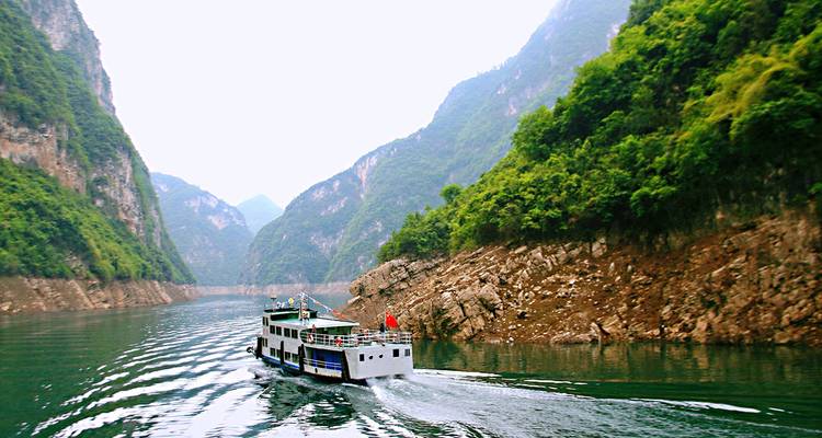 Petit bateau de passagers naviguant à travers une gorge verte escarpée sur le fleuve Yangtsé laissant un sillage ondulant derrière lui.