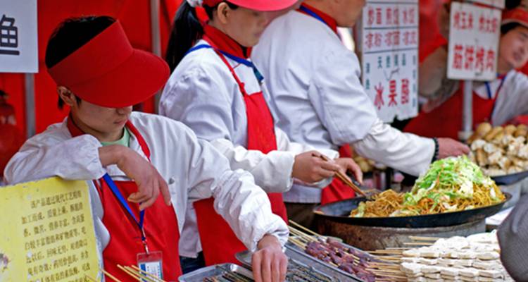 Vendedores de comida callejera con uniformes blancos y gorras rojas preparan brochetas y fideos en un puesto bullicioso.