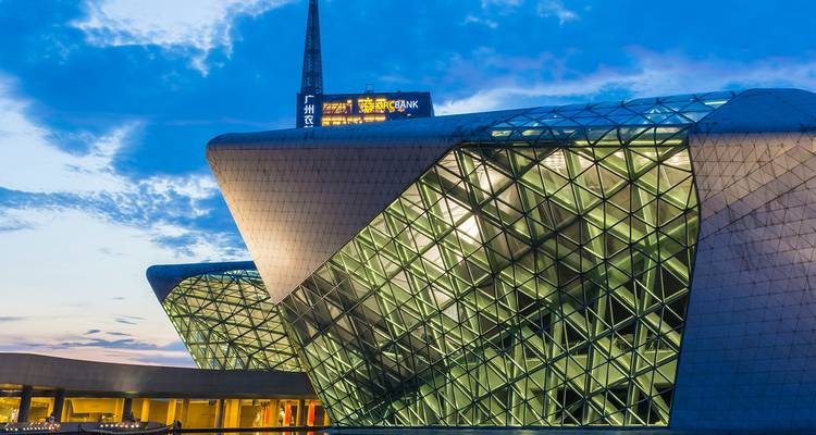 Hoekige glazen gevel van het Guangzhou Opera House verlicht in de schemering onder een blauw-gouden hemel.