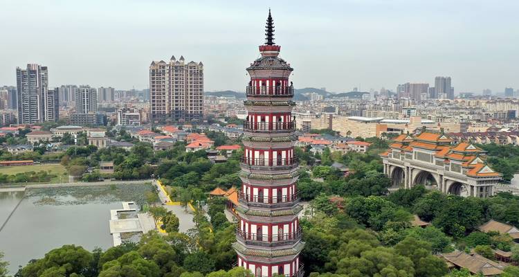 Een hoge rood-wit gestreepte pagode rijst op boven een weelderig stadspark met een moderne Chinese stadshorizon erachter.