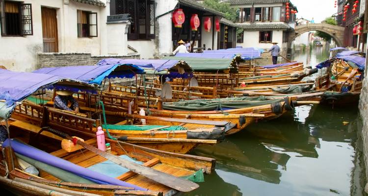 Reihe von hölzernen Sampan-Booten mit lila Überdachungen, die entlang eines schmalen Kanals vertäut sind, der von weiß getünchten traditionellen Häusern gesäumt wird.