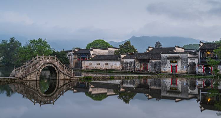 Ancient stone bridge and traditional Chinese village reflected perfectly in a calm lake at dawn.