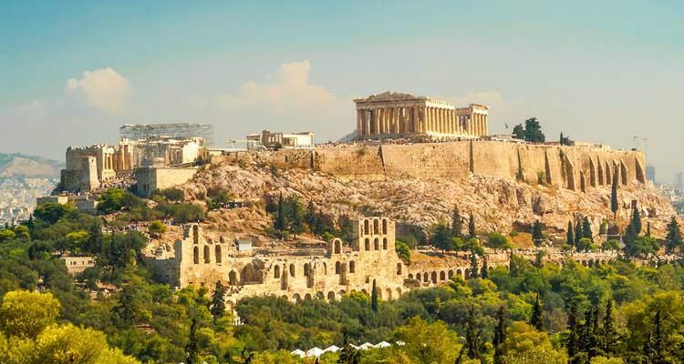 Vue panoramique de l'Acropole d'Athènes perchée au sommet d'une colline rocheuse entourée d'arbres verts et d'un paysage urbain sous une lumière chaleureuse.