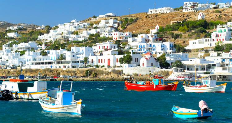 Des maisons cycladiques blanchies à la chaux dégringolent le long d'une colline au-dessus d'une baie remplie de bateaux de pêche colorés à Mykonos.