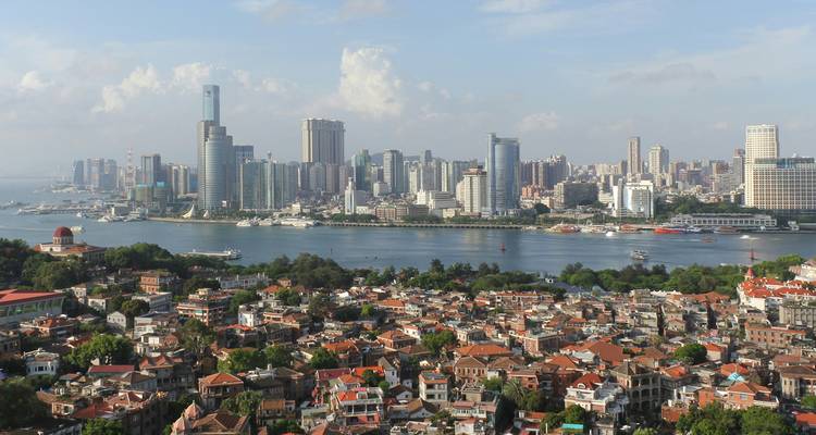 Skyline van Xiamen met moderne hoogbouw aan de overkant van een brede rivier, gezien vanaf een verhoogd uitkijkpunt boven pannendaken.