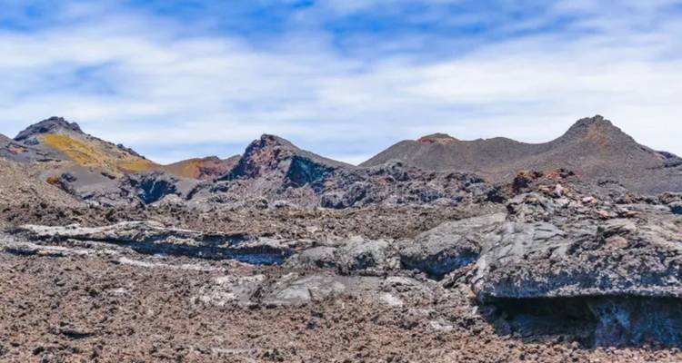 Paisaje volcánico accidentado con rocas de lava oscuras y conos de ceniza distantes bajo un cielo azul.