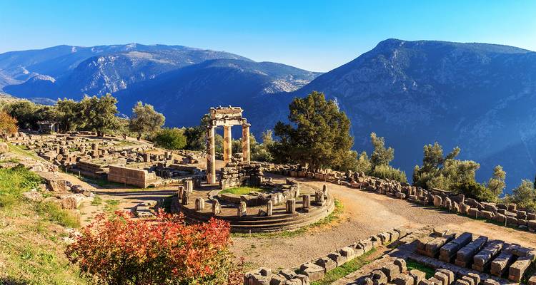 Ruinas circulares del templo antiguo de Delfos enmarcadas por montañas y un cielo azul brillante.