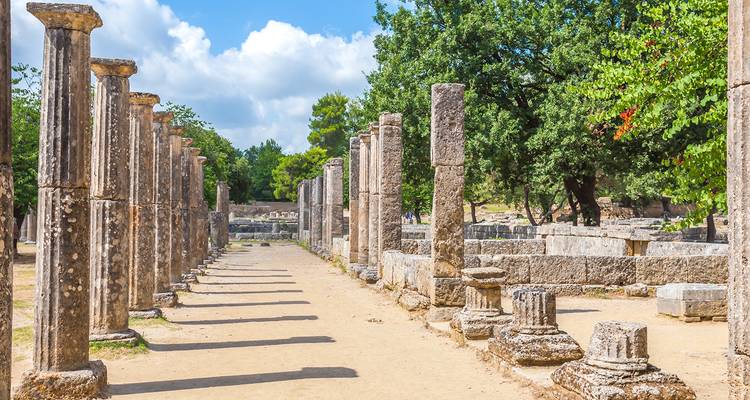 Colonnade ensoleillée et ruines de l'Olympie antique avec des arbres et un ciel bleu.