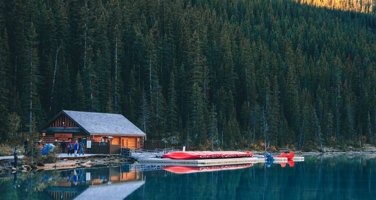 Des canoës colorés reposent près d'un cabanon rustique au bord du lac, entourés d'une dense forêt de conifères qui se reflète dans les eaux alpines calmes au crépuscule.
