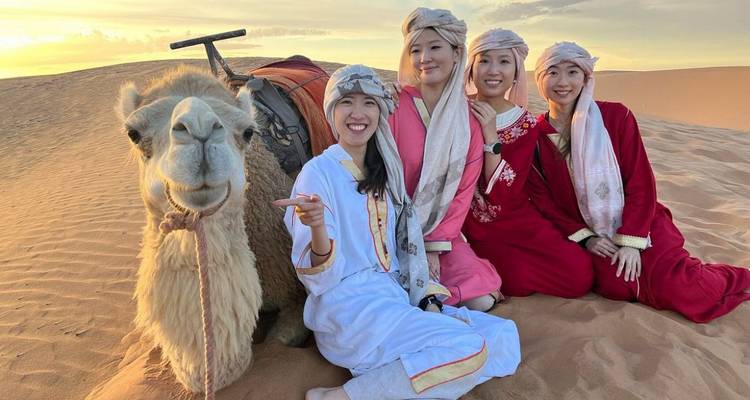 Cuatro mujeres con túnicas tradicionales del desierto posando con un camello amigable en dunas doradas al atardecer.