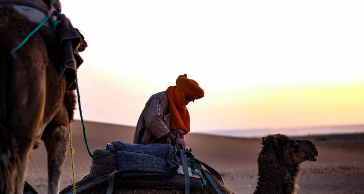 Guía bereber con turbante naranja ajustando el equipo del camello al amanecer en las dunas.