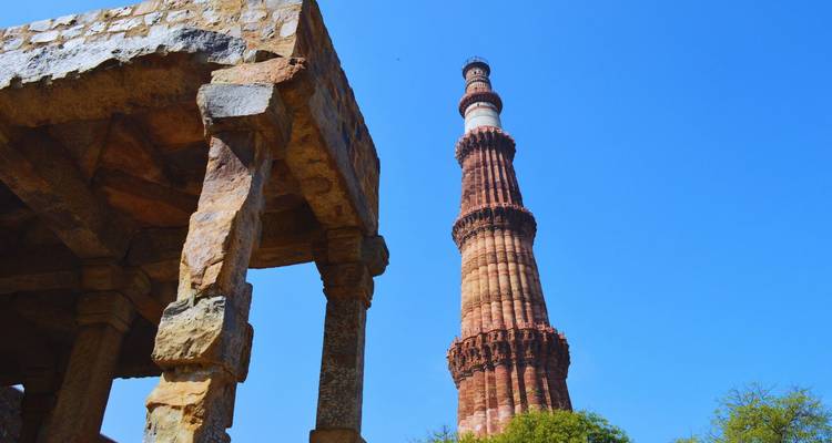 Der aufragende rote Sandstein Qutub Minar gerahmt von antiken Steinruinen unter einem klaren blauen Himmel.