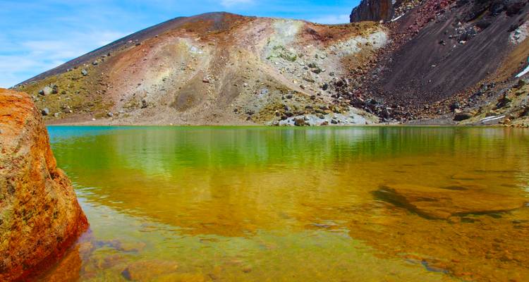 Ein mineralreicher Alpensee mit lebendigen grünen, gelben und rostigen Farbtönen unterhalb felsiger Hänge.