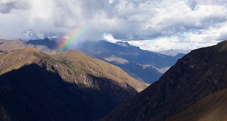 Bergtalszene mit dramatischen Wolken und einem Regenbogen, der entfernte Gipfel beleuchtet.