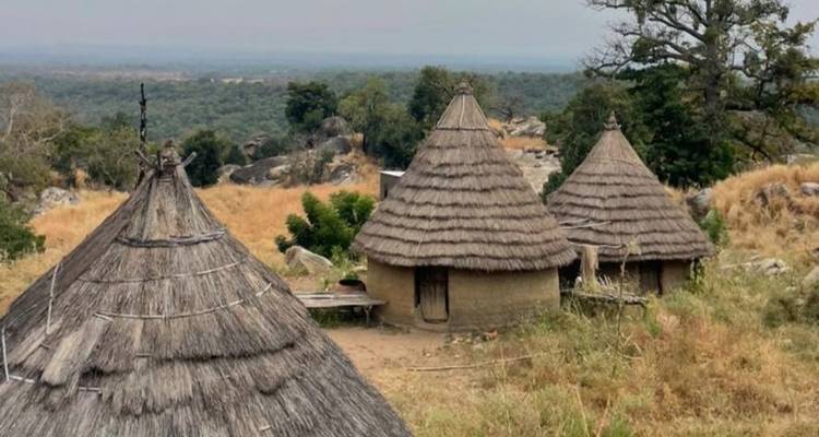 Vue panoramique de huttes circulaires au toit de chaume dans un paysage rural de savane avec des collines au loin.