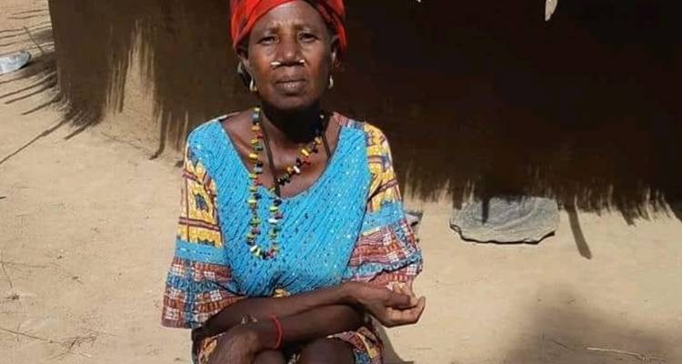 Portrait d'une femme âgée en robe bleue à motifs et foulard rouge assis devant une hutte dans une cour poussiéreuse.