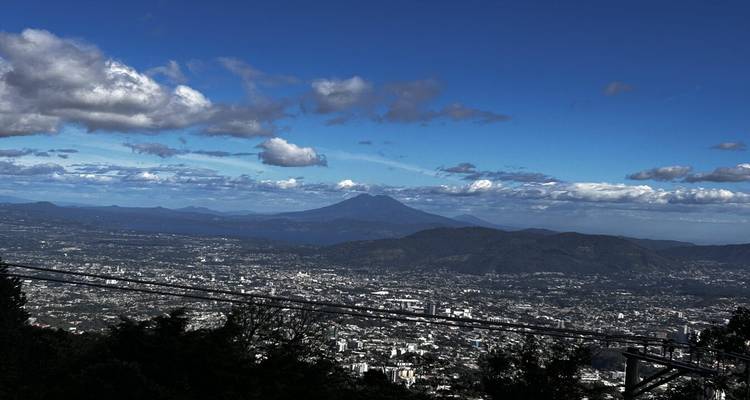 Vue panoramique depuis un belvédère de montagne sur la vallée de San Salvador avec paysage urbain et volcans lointains sous un ciel bleu.