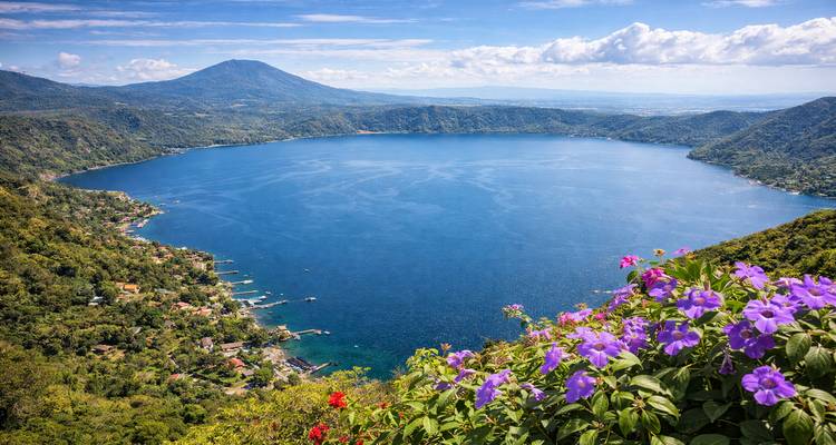 Lac de cratère Coatepeque d'un bleu éclatant entouré de collines vertes avec des fleurs violettes au premier plan.