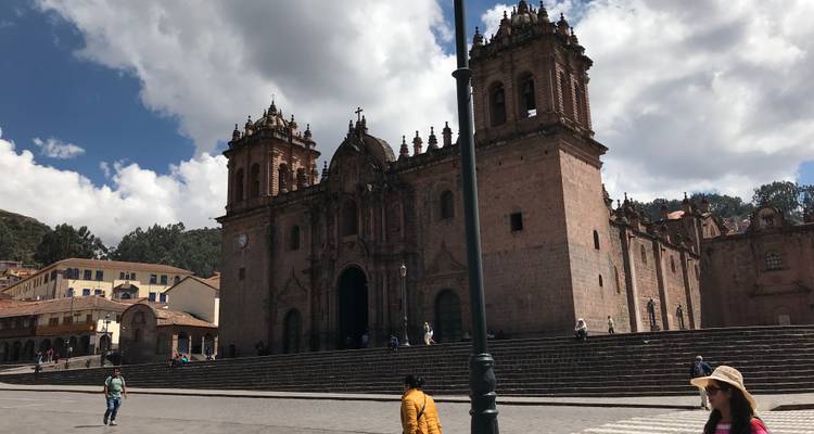 Historic Cusco Cathedral towering over wide stone steps with scattered visitors under partly cloudy sky.