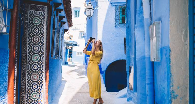 Vrouw in een wapperende gele jurk fotografeert de levendige blauwe steegjes van Chefchaouen met een kleine camera.
