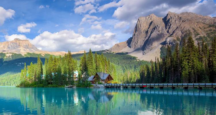 Cabane idyllique sur une île boisée se reflétant dans les eaux vert émeraude d'un lac avec des sommets imposants des montagnes Rocheuses sous un ciel bleu parsemé de nuages.