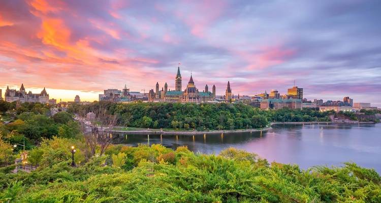 Dramatic sunset over a riverside parliament complex surrounded by lush trees and water.