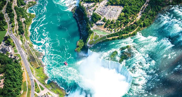 Aerial of massive horseshoe waterfall with turquoise rapids and a tour boat at its base.