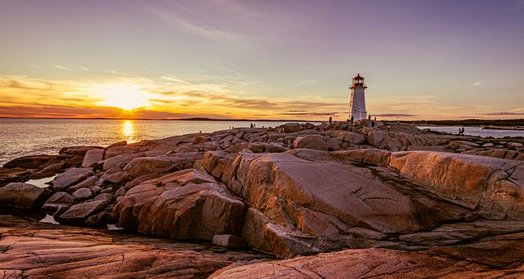 Classic white lighthouse atop rugged granite rocks during a colourful ocean sunset.
