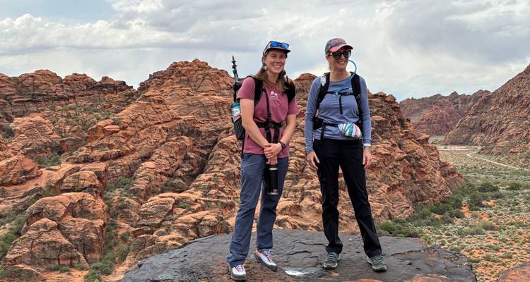 Zwei Wanderer stehen auf dunklem Vulkangestein mit dramatischen roten Sandsteinformationen dahinter unter einem teilweise bewölkten Himmel.