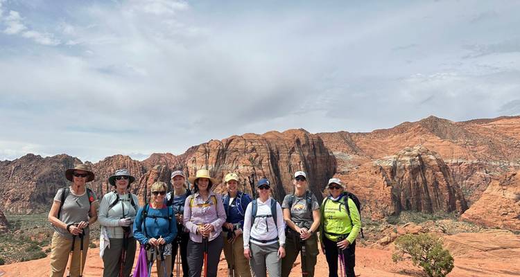 Gruppe von Wanderern posiert auf rotem Sandsteinplateau mit schroffen Snow Canyon Klippen dahinter und bewölktem Himmel darüber.