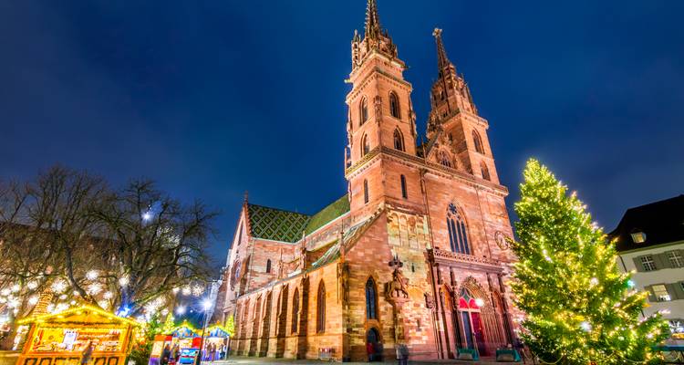 Cathédrale de Bâle et stands de marché de Noël festifs illuminés à côté d'un arbre imposant la nuit.