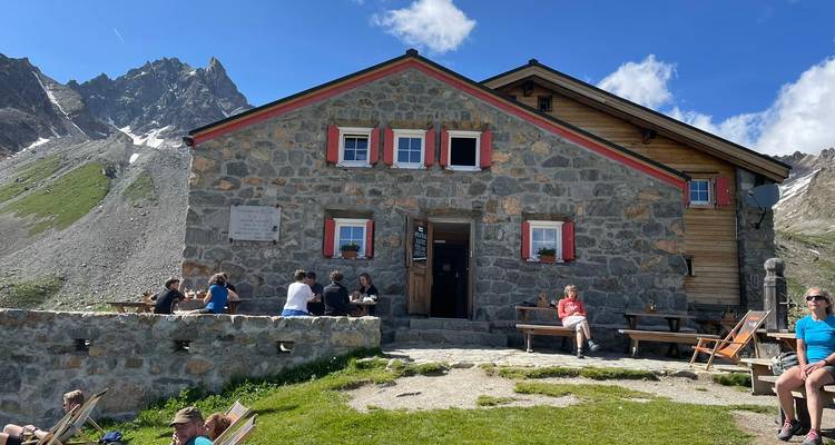 Refuge de montagne en pierre avec des randonneurs se reposant aux tables de pique-nique sous un ciel bleu éclatant.