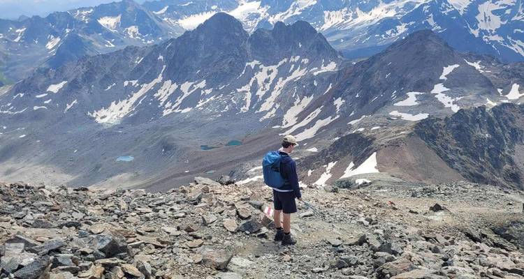 Un randonneur avec un sac à dos se tient sur une crête rocheuse de montagne surplombant des pics alpins enneigés spectaculaires et de petits lacs de montagne turquoise en contrebas.