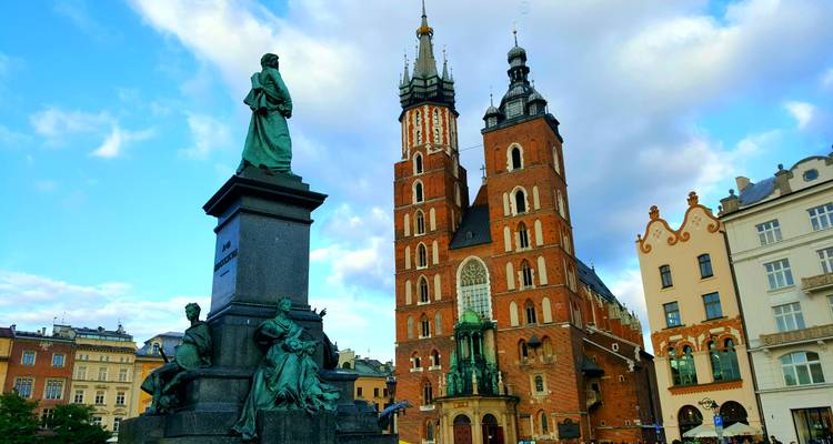 La Basílica de Santa María y las estatuas de bronce dominan la plaza principal iluminada por el sol de Cracovia bajo cielos azules con nubes dispersas