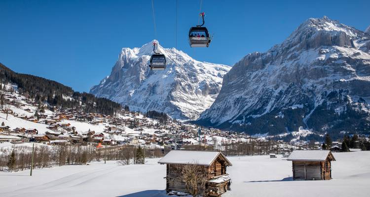 Paysage alpin enneigé avec deux téléphériques glissant au-dessus de chalets en bois et un village niché sous des montagnes imposantes.